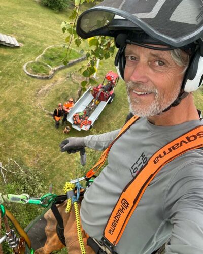 En man som tar en selfie på sig själv uppe från ett träd som han håller på att fälla. På marken ser man hans kollegor.